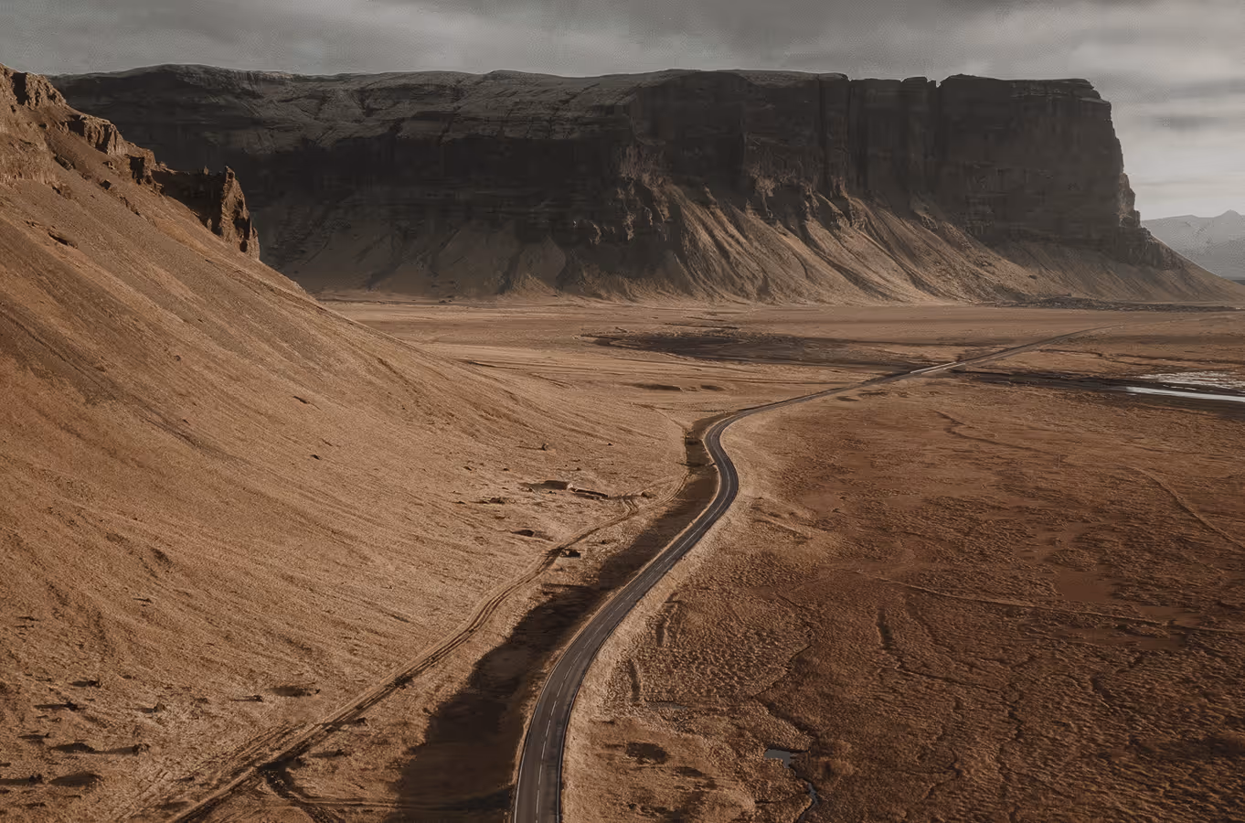 Winding road through a barren, rocky desert landscape with steep cliffs in the background under a cloudy sky.