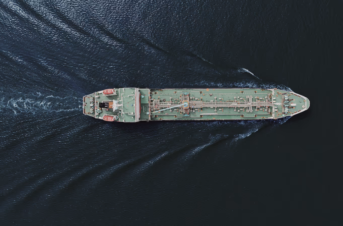 Aerial view of a large cargo ship sailing through dark water, leaving a V-shaped wake behind.